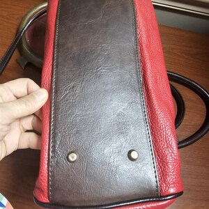 May include: The bottom of a handbag featuring a rectangular dark brown panel with a textured finish, surrounded by bright red pebbled sides. Four silver-toned metal feet are attached to the corners of the brown base to provide stability.