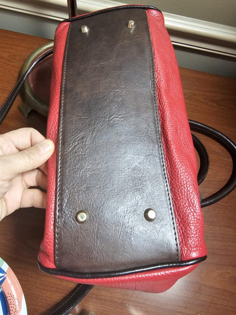 May include: The bottom of a handbag featuring a rectangular dark brown panel with a textured finish, surrounded by bright red pebbled sides. Four silver-toned metal feet are attached to the corners of the brown base to provide stability.