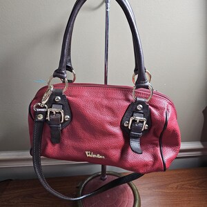 May include: A red textured handbag with dark brown leather handles and trim. The bag features gold-tone hardware, including two decorative buckles on the front, a top zipper closure, and the brand name Valentina printed in gold lettering on the front.