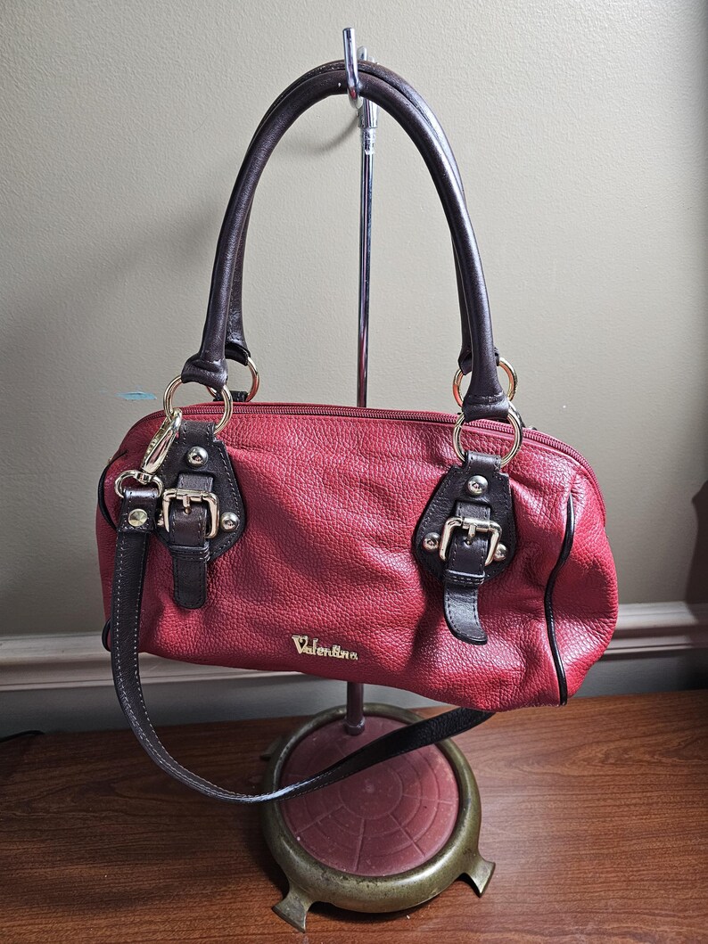 May include: A red textured handbag with dark brown leather handles and trim. The bag features gold-tone hardware, including two decorative buckles on the front, a top zipper closure, and the brand name Valentina printed in gold lettering on the front.