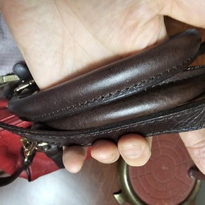 May include: A dark brown leather handbag handle with visible stitching, featuring a smooth, rounded top section and a flat, textured strap attached to the side, with a glimpse of a red leather bag body visible underneath.