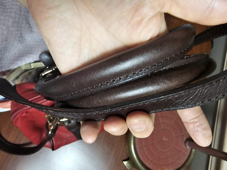 May include: A dark brown leather handbag handle with visible stitching, featuring a smooth, rounded top section and a flat, textured strap attached to the side, with a glimpse of a red leather bag body visible underneath.