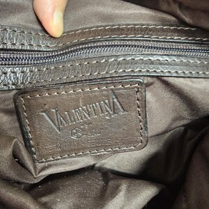 May include: A dark brown interior lining of a bag featuring a rectangular leather patch embossed with the text VALENTINA. The patch includes a small logo and the words MADE IN ITALY. A zippered pocket with a dark brown zipper pull is visible above.