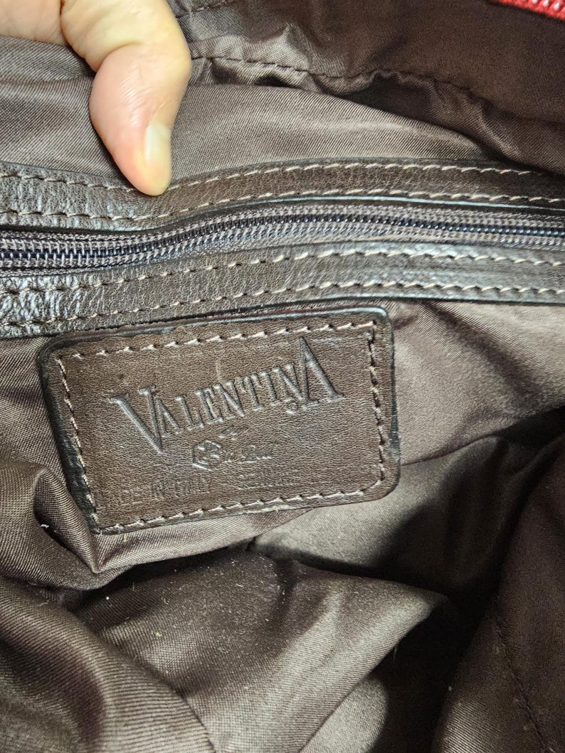 May include: A dark brown interior lining of a bag featuring a rectangular leather patch embossed with the text VALENTINA. The patch includes a small logo and the words MADE IN ITALY. A zippered pocket with a dark brown zipper pull is visible above.