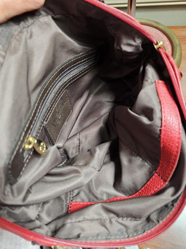 May include: A red handbag with a textured exterior and a dark brown fabric interior lining. The interior features a zippered pocket with a gold-toned metal pull and a rectangular leather patch embossed with the brand name Luv Betsey.