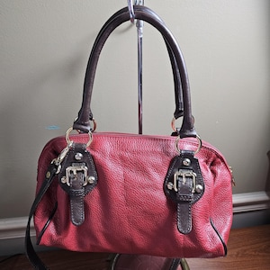 May include: A red textured handbag with dark brown leather handles and trim. The bag features two decorative buckle accents on the front, gold-tone metal hardware, and a top zipper closure. It includes a detachable dark brown shoulder strap.