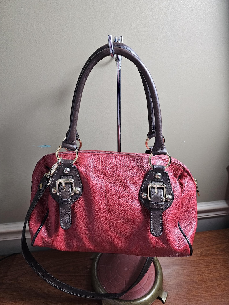May include: A red textured handbag with dark brown leather handles and trim. The bag features two decorative buckle accents on the front, gold-tone metal hardware, and a top zipper closure. It includes a detachable dark brown shoulder strap.