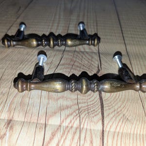 May include: Two antique-style cabinet handles. The handles are made of bronze-colored metal with a decorative design. Each handle has two mounting posts with silver-colored heads. The handles are on a wooden surface.