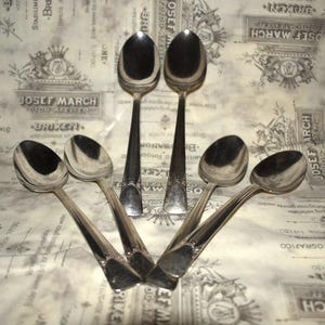 May include: Five silver spoons with ornate handles. The spoons are arranged in a fan shape on a white background with a pattern of black and gray text.