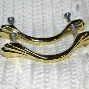 May include: Two gold-toned cabinet door pulls with a decorative, ornate design. The pulls have a curved shape and are attached with screws.