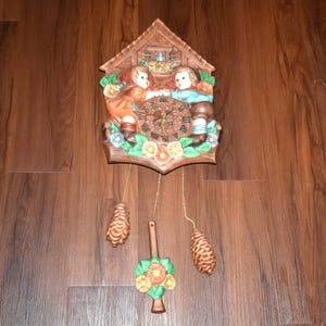 May include: A brown wall clock shaped like a house with a central circular dial featuring Roman numerals. Two figures in orange and blue clothing stand on either side of the dial, surrounded by painted flowers. Three hanging weights are attached.