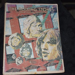 May include: Vintage songbook titled "The Rolling Stones" featuring portraits of the band members. The cover has a red and blue background with images of the band members' faces. The book is slightly worn, suggesting its age.