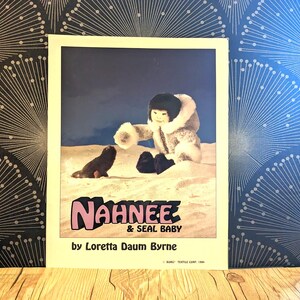 May include: A book cover featuring a child in a white and brown hooded parka, sitting in the snow with a seal pup. The book title is "Nahnee & Seal Baby" by Loretta Daum Byrne.
