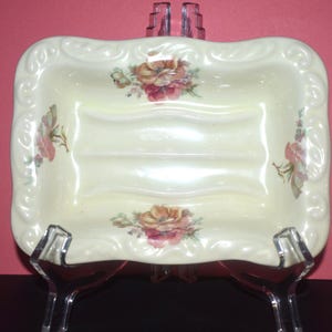 May include: A rectangular cream-colored ceramic soap dish featuring a raised, scalloped border and a central floral pattern with pink and orange roses. The dish has two horizontal ridges in the center and is displayed on a clear plastic stand.