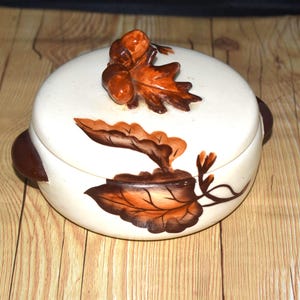 May include: A white ceramic dish with a brown lid. The dish is decorated with brown and orange leaves and acorns.