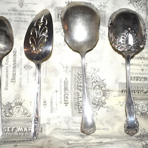 May include: Four silver serving spoons with ornate designs. The spoons have a polished finish and are arranged on a white and gray patterned surface.