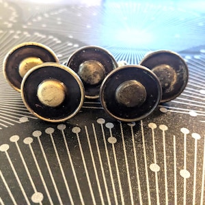 May include: A set of four circular buttons featuring a dark black center with a raised, metallic gold-toned rim and a matching gold-toned circular accent in the middle. Each button has a smooth, polished finish and a distinct, layered design.