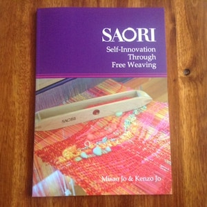 May include: A book titled "SAORI Self-Innovation Through Free Weaving" with a purple and white cover. The book features a colorful woven textile and a wooden weaving tool. The authors' names, Misae Jo & Kenzo Jo, are printed at the bottom.