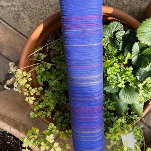 K&ouml;nnte beinhalten: Eine Rolle blau-violettes Baumwollgarn mit horizontalen Streifen in verschiedenen Farben. Das Garn ist mit japanischem Text und Ma&szlig;en versehen. Die Rolle befindet sich vor einem Terrakotta-Topf mit gr&uuml;nen Pflanzen.