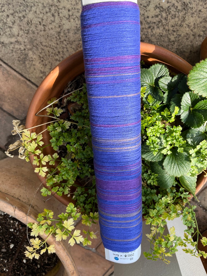 K&ouml;nnte beinhalten: Eine Rolle blau-violettes Baumwollgarn mit horizontalen Streifen in verschiedenen Farben. Das Garn ist mit japanischem Text und Ma&szlig;en versehen. Die Rolle befindet sich vor einem Terrakotta-Topf mit gr&uuml;nen Pflanzen.