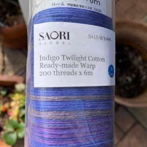 K&ouml;nnte beinhalten: Eine Rolle SAORI GLOBAL Indigo Twilight Cotton Fertig-Webkette. Das Etikett lautet "200 F&auml;den x 6m". Die Fadenfarben sind Blaut&ouml;ne, Lila und Grau. Das Produkt ist in klarem Kunststoff verpackt.