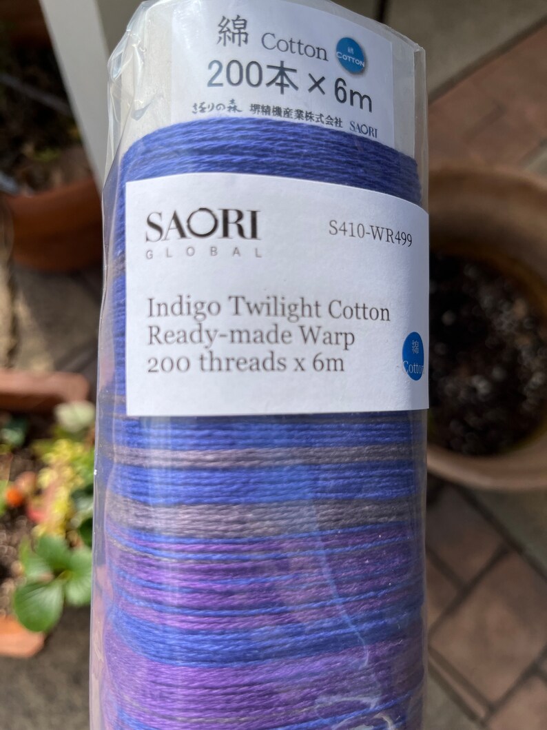 K&ouml;nnte beinhalten: Eine Rolle SAORI GLOBAL Indigo Twilight Cotton Fertig-Webkette. Das Etikett lautet "200 F&auml;den x 6m". Die Fadenfarben sind Blaut&ouml;ne, Lila und Grau. Das Produkt ist in klarem Kunststoff verpackt.