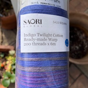 K&ouml;nnte beinhalten: Eine Rolle Indigo Twilight Baumwoll-Fertigschussfaden. Das Etikett lautet "SAORI GLOBAL" und "200 F&auml;den x 6m". Der Faden ist eine Mischung aus blauen, violetten und grauen Farbt&ouml;nen. Die Verpackung ist aus klarem Kunststoff.