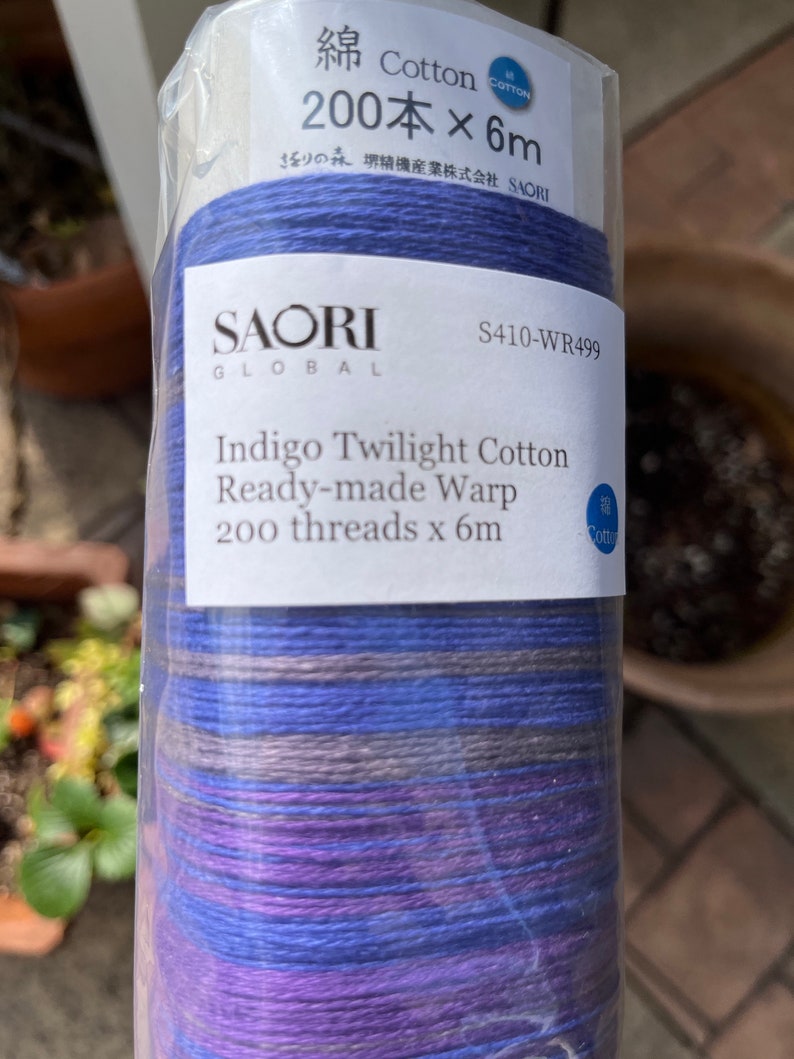 K&ouml;nnte beinhalten: Eine Rolle Indigo Twilight Baumwoll-Fertigschussfaden. Das Etikett lautet "SAORI GLOBAL" und "200 F&auml;den x 6m". Der Faden ist eine Mischung aus blauen, violetten und grauen Farbt&ouml;nen. Die Verpackung ist aus klarem Kunststoff.