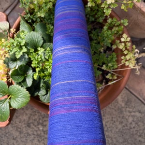 K&ouml;nnte beinhalten: Eine hohe Spule aus blauem Garn mit horizontalen Streifen in Lila, Rosa und Gold. Das Garn befindet sich in einem Topf mit gr&uuml;nen Pflanzen. Das Garn ist wahrscheinlich f&uuml;r Strick- oder H&auml;kelprojekte.