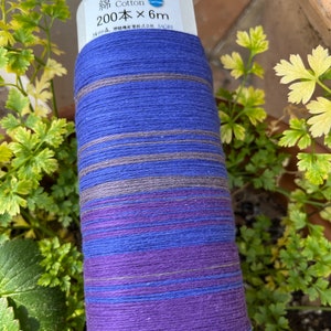 K&ouml;nnte beinhalten: Eine Rolle blaues, violettes und graues Baumwollgarn. Auf dem Etikett steht "Cotton 200 x 6m". Das Garn ist um eine wei&szlig;e Papprolle gewickelt. Gr&uuml;ne Pflanzen sind im Hintergrund.