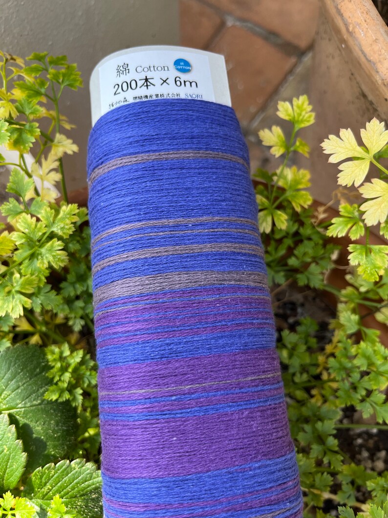 K&ouml;nnte beinhalten: Eine Rolle blaues, violettes und graues Baumwollgarn. Auf dem Etikett steht "Cotton 200 x 6m". Das Garn ist um eine wei&szlig;e Papprolle gewickelt. Gr&uuml;ne Pflanzen sind im Hintergrund.