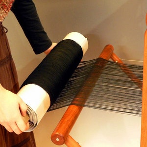 May include: A wooden weaving loom with black thread being wound onto a large spool. The spool is held by a person, and the loom's frame is made of light brown wood. The black thread is stretched across the loom, ready for weaving.