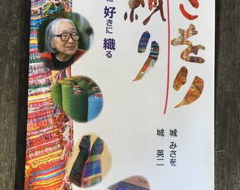 May include: A book cover featuring the Japanese title "さをり" in red and orange, with the text "好きに好きに織る". The cover includes a portrait of a woman and colorful woven textile samples. The book is on a wooden surface.