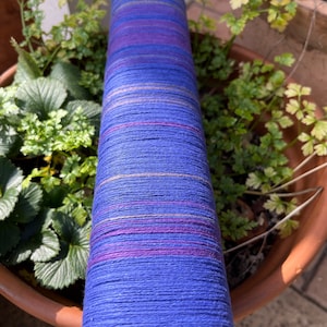 K&ouml;nnte beinhalten: Eine zylindrische Spule aus blauem Garn mit horizontalen Streifen in Lila, Rosa und Gold. Das Garn ist eng gewickelt und hat eine strukturierte Oberfl&auml;che. Die Spule steht vertikal, mit gr&uuml;nen Pflanzen im Hintergrund.