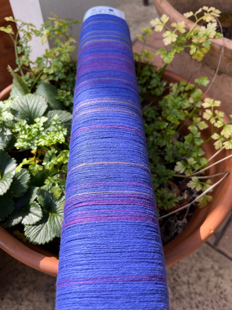 K&ouml;nnte beinhalten: Eine zylindrische Spule aus blauem Garn mit horizontalen Streifen in Lila, Rosa und Gold. Das Garn ist eng gewickelt und hat eine strukturierte Oberfl&auml;che. Die Spule steht vertikal, mit gr&uuml;nen Pflanzen im Hintergrund.