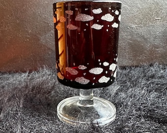 May include: A dark brown glass goblet featuring a repeating pattern of small, textured silver clouds encircling the exterior. The vessel is supported by a clear, circular glass pedestal base, creating a distinct contrast in color and transparency.