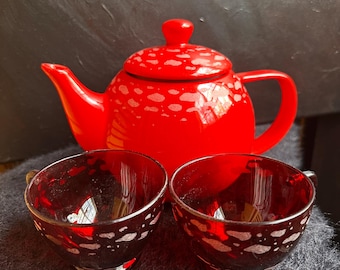 May include: A bright red ceramic teapot with a matching lid and two dark red glass teacups. The teapot features a light-colored speckled pattern near the top, while the two glass teacups have a similar speckled design around their exterior.