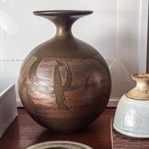 May include: A brown ceramic vase with a wide, round body and a narrow neck. The vase has a unique, abstract design with brown and orange brushstrokes.