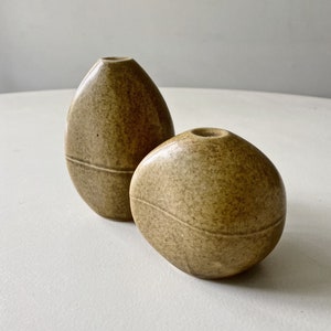 May include: Two small, speckled, light brown ceramic vases shaped like rounded stones. The vases have a subtle, horizontal line around the middle.