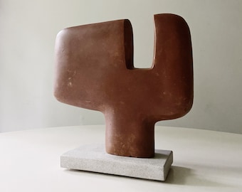 May include: A terracotta-colored abstract sculpture with a rectangular base. The sculpture has a rounded, geometric shape with a cutout section. The base is a light gray, rectangular stone. The sculpture is displayed on a white surface.