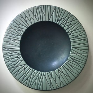 May include: A dark green ceramic plate with a geometric design around the rim. The plate has a raised, textured pattern of lines that radiate outwards from the centre.