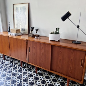 May include: A mid-century modern teak wood sideboard with sliding doors and three cabinet doors. The sideboard is sitting on a black and white patterned tile floor. There is a black lamp with a silver arm on the top of the sideboard. There are also a few decorative items on top of the sideboard, including a potted plant, a framed print, and a few sculptures.