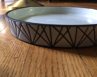May include: A white ceramic tray with a black geometric design around the rim. The tray is sitting on a wooden surface.