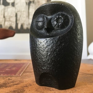 May include: A black cast iron owl figurine with a detailed face and closed eyes. The owl is sitting on a brown surface.