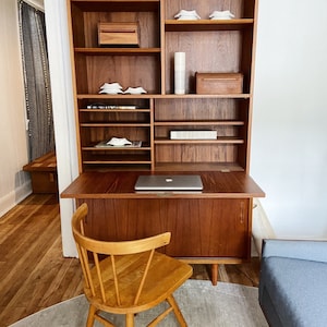 May include: A wooden desk with a drop-down front and a built-in bookcase. The desk has a light wood finish and a chair with a natural wood finish. The bookcase has multiple shelves and is filled with books and decorative items.