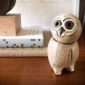 May include: A ceramic owl figurine with a round head and body, featuring dark brown eyes and beak. The owl is light beige with darker brown accents and stands on three small feet. The figurine is on a wooden surface.