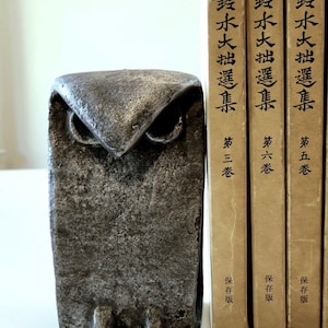 May include: A gray metal owl bookend with a textured surface. The bookend is sitting in front of three books with Japanese text on the spines. The text on the spines reads "鈴木大拙選集" and "保存版".