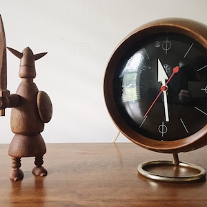 Puede incluir: Un reloj de madera con una esfera negra y agujas blancas. El reloj tiene forma de esfera y está sobre un soporte dorado. Una figura de madera de un guerrero con una espada está de pie a la izquierda del reloj.