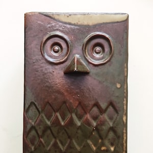 May include: A ceramic owl sculpture with a brown, green and white glaze. The owl has large, round eyes and a diamond-shaped pattern on its body.