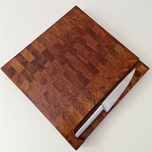 Puede incluir: Una tabla de cortar grande, rectangular y de madera con una ranura para cuchillo. La tabla es de madera oscura con un patrón de tablero de ajedrez. El cuchillo es plateado y tiene un mango negro.